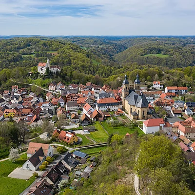 Luftaufnahme einer kleinen Stadt mit einer großen Kirche und einem Schloss, umgeben von bewaldeten Hügeln.
