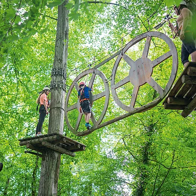 Drei Personen mit Kletterausrüstung auf einem Hochseilgarten-Parcours zwischen Bäumen und Holzplattformen.