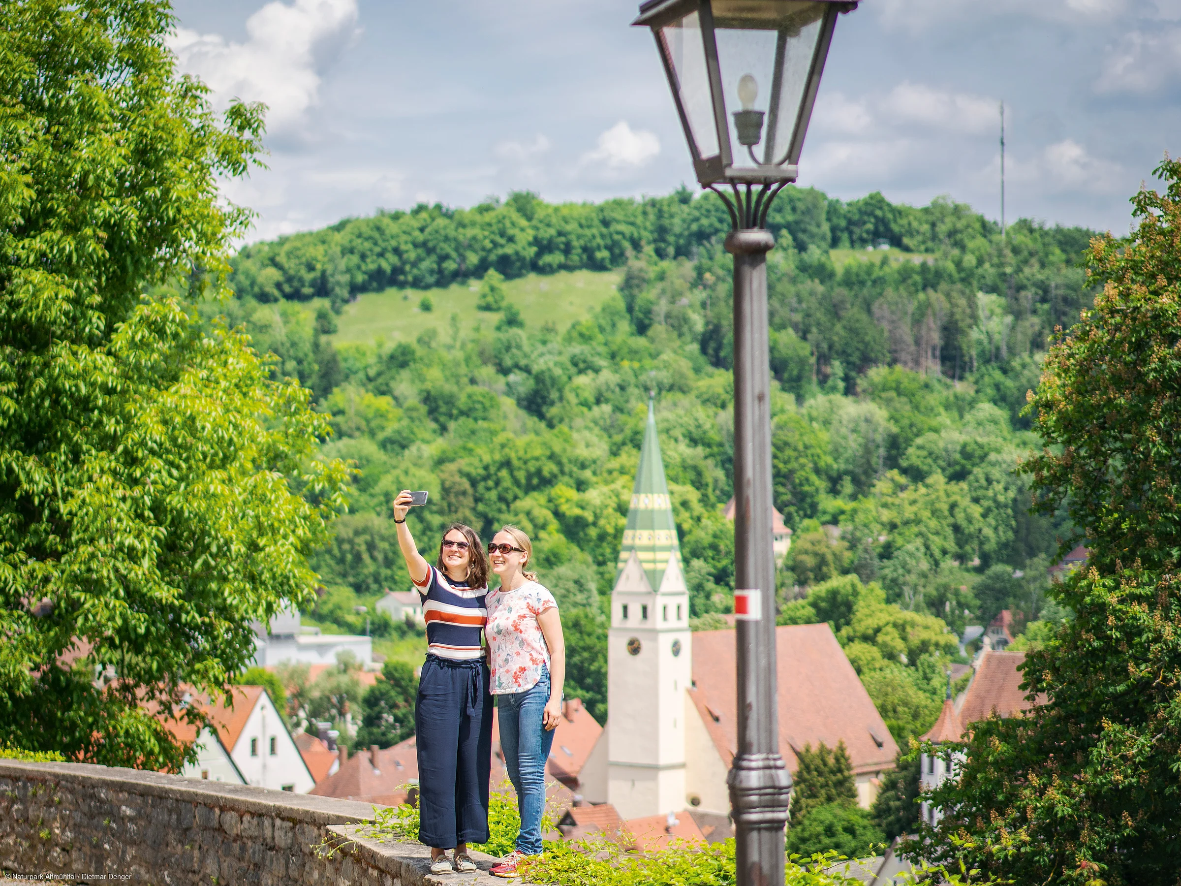 Zwei Frauen machen Selfie vor grünem Hügel und Kirche mit spitzem Turm, Laterne im Vordergrund.