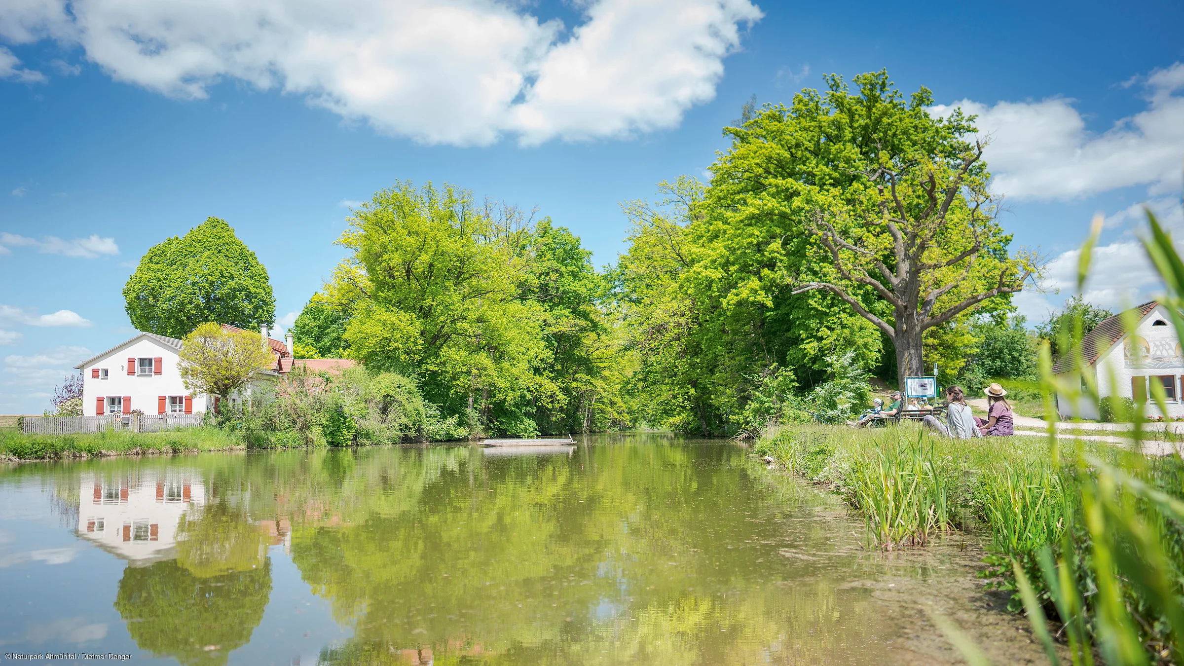 Fluss mit grünen Bäumen, zwei Häusern links und rechts und Personen am Ufer unter blauem Himmel