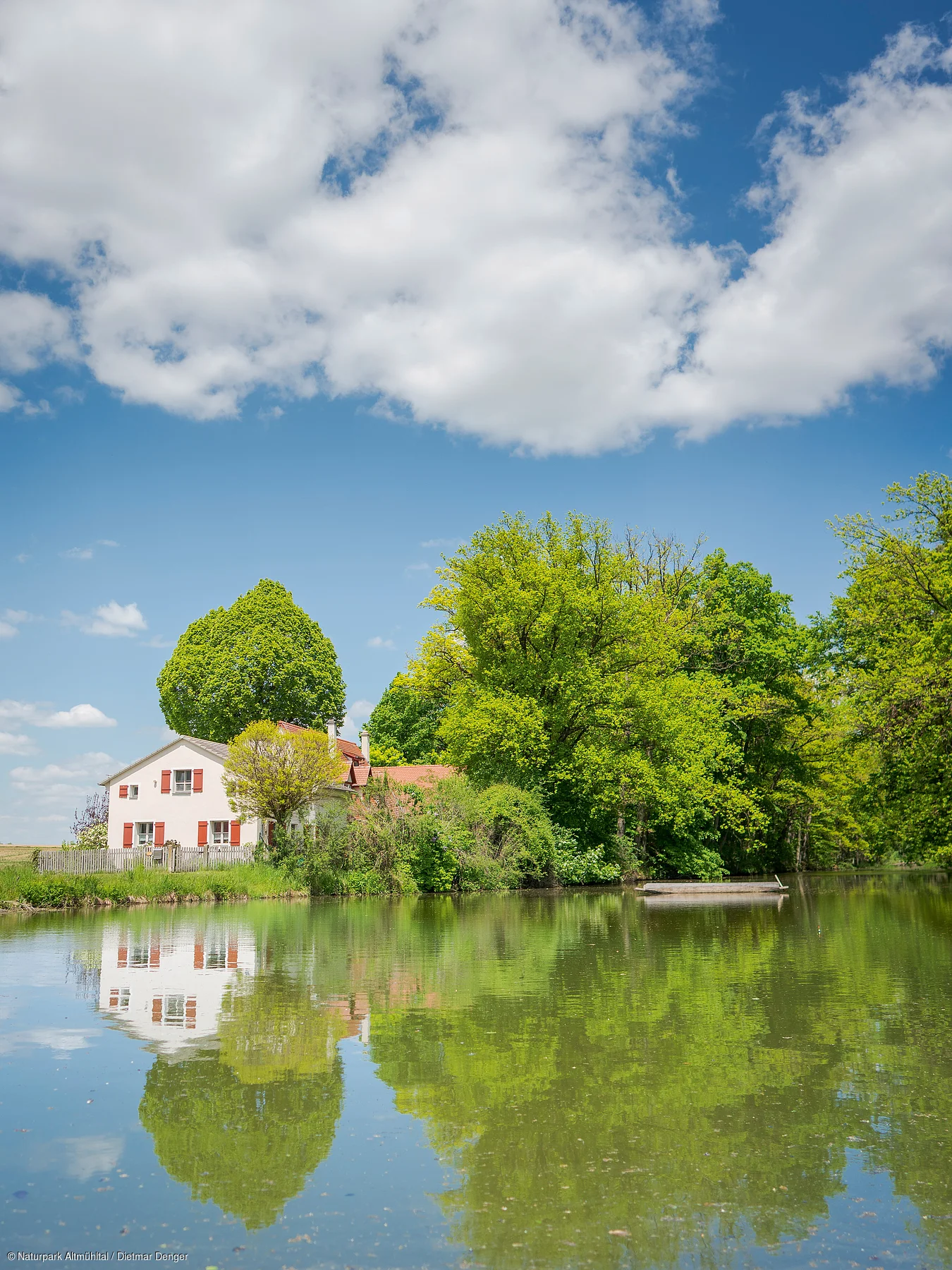 Haus mit roten Fensterläden am Ufer eines Sees, umgeben von grünen Bäumen und blauem Himmel mit Wolken