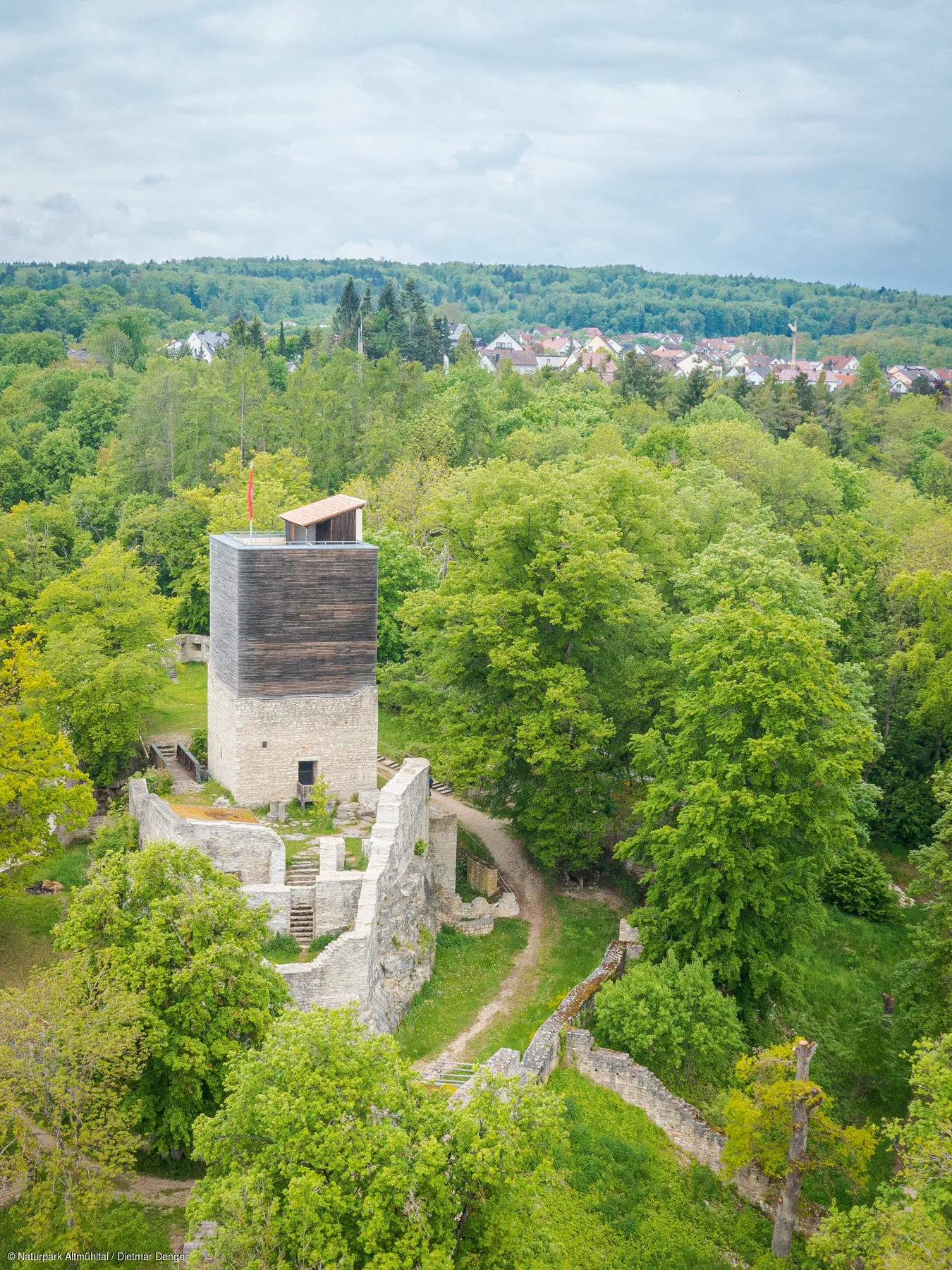 Burgruine mit Turm und umgebender Mauer in einem bewaldeten Gebiet nahe eines Dorfes unter bewölktem Himmel.