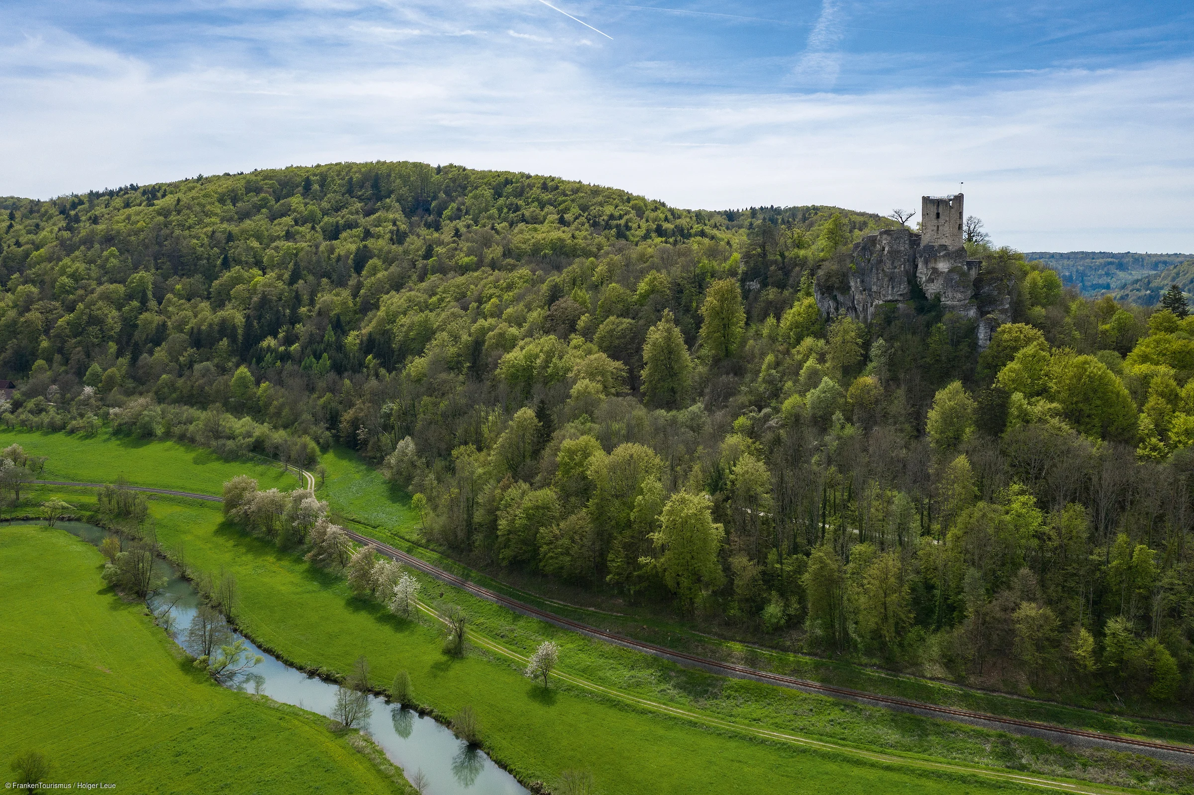 Blick auf bewaldeten Hügel mit Burgruine, Fluss und grünen Wiesen unter blauem Himmel mit Wolken.