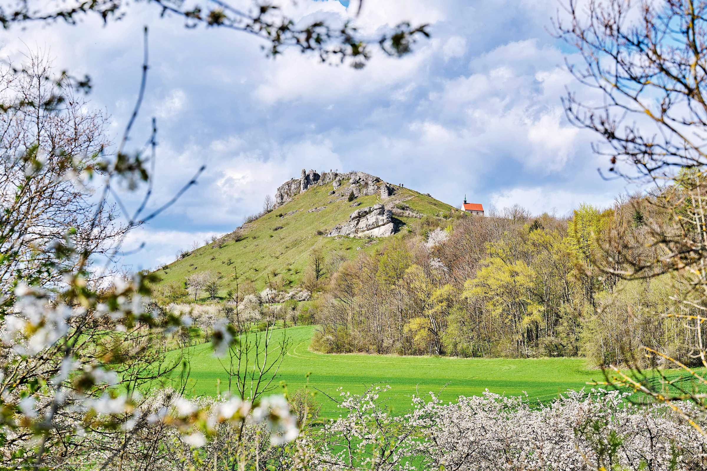 Grüner Berg mit Felsen und Kapelle, umgeben von blühenden Bäumen und bewölktem Himmel.