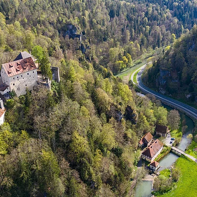 Luftaufnahme einer Burg auf bewaldetem Hügel neben einem Fluss und einer kurvigen Straße im Tal.