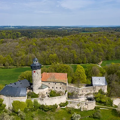 Luftaufnahme einer historischen Burg mit Turm, umgeben von Wald und Feldern unter blauem Himmel.