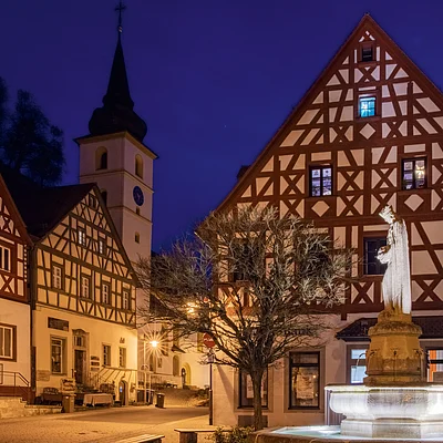 Fachwerkhäuser und beleuchteter Brunnen mit Statue in Ortsmitte bei Nacht, Kirche im Hintergrund.