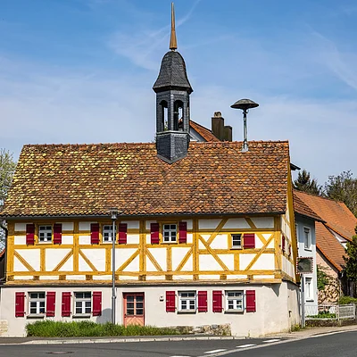 Fachwerkhaus mit rotem Schindeldach und kleinem Turm an einer Straßenecke, umgeben von Bäumen.