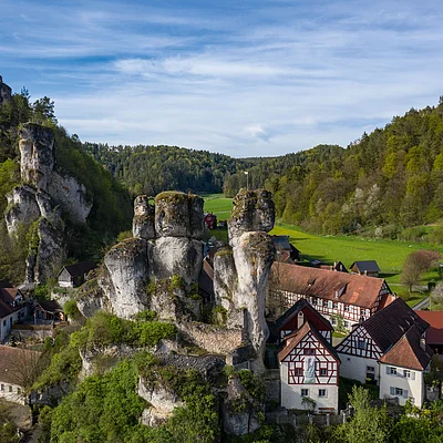 Dorf mit Fachwerkhäusern und Felsen umgeben von bewaldeten Hügeln unter blauem Himmel