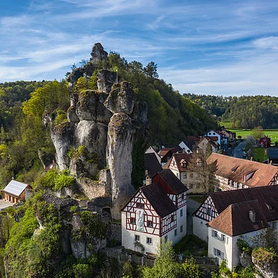 Felsenformation mit Fachwerkhäusern und Wald im Hintergrund unter blauem Himmel mit Wolken.
