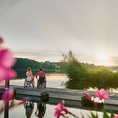 Zwei Personen im Rollstuhl sitzen auf einem Steg am See, Sonnenuntergang und Blumen im Vordergrund.