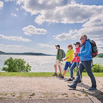 Vier Personen mit Rucksäcken wandern auf einem Weg entlang eines Sees bei bewölktem Himmel.