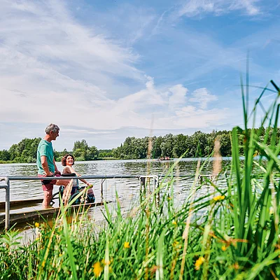 Mann und Frau mit Rollstuhl am Seeufer auf Steg mit Wasser und Bäumen im Hintergrund bei sonnigem Himmel