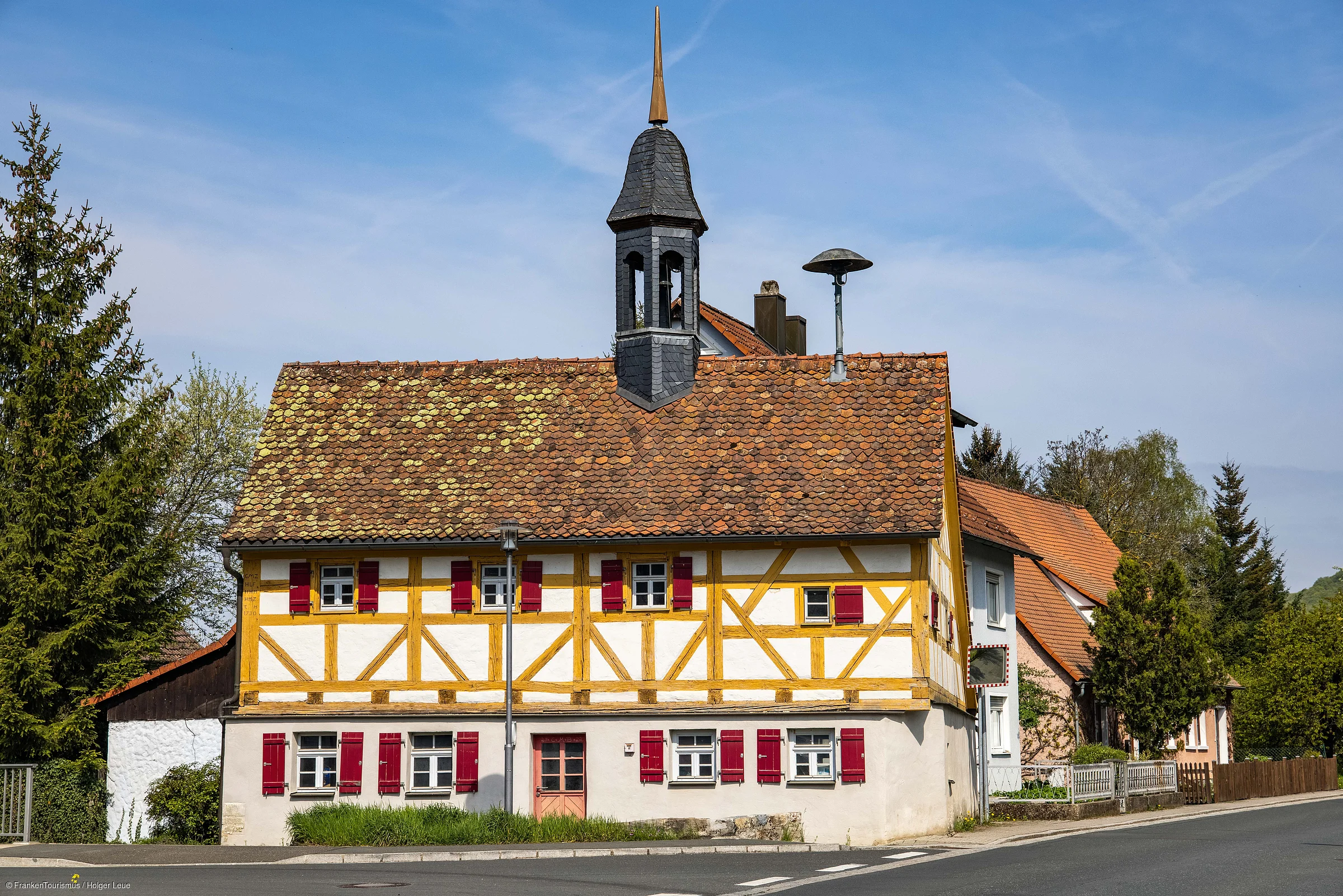 Fachwerkhaus mit rotem Schindeldach und kleinem Turm an einer Straßenecke, umgeben von Bäumen.
