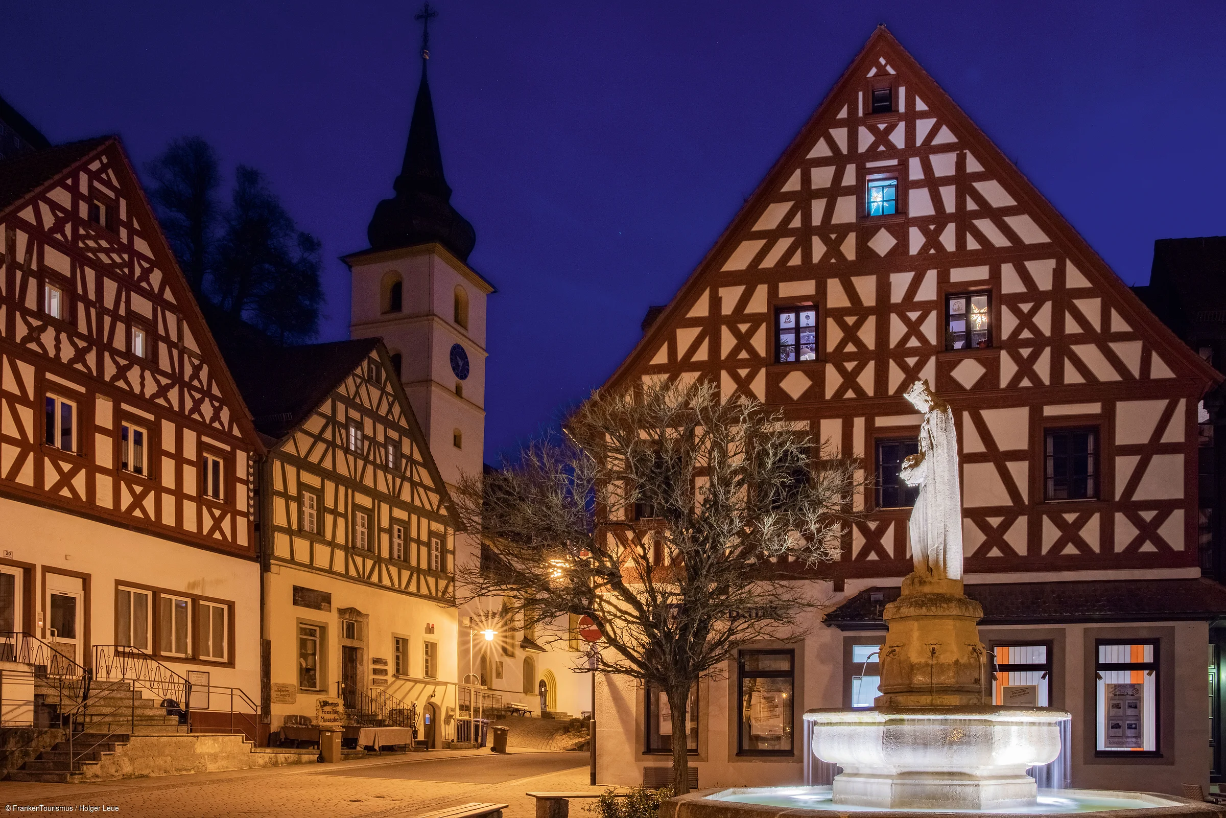 Fachwerkhäuser und beleuchteter Brunnen mit Statue in Ortsmitte bei Nacht, Kirche im Hintergrund.