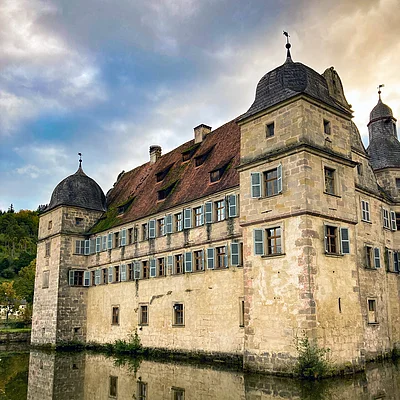 Historisches Schloss mit Türmen und blauen Fensterläden, Spiegelung im Wasser vor bewölktem Himmel