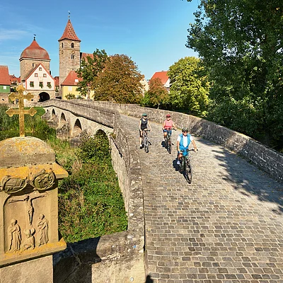 Drei Radfahrer fahren auf einer gepflasterten Steinbrücke mit historischen Gebäuden und Bäumen im Hintergrund.