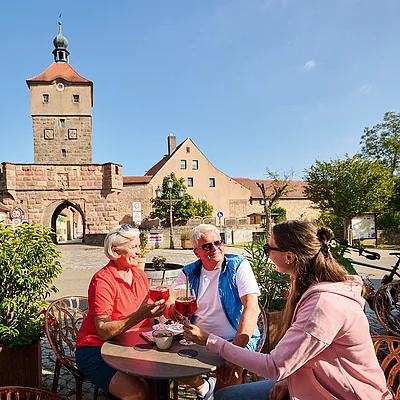 Drei Personen sitzen draußen an einem Tisch vor historischem Stadttor und stoßen mit Getränken an.