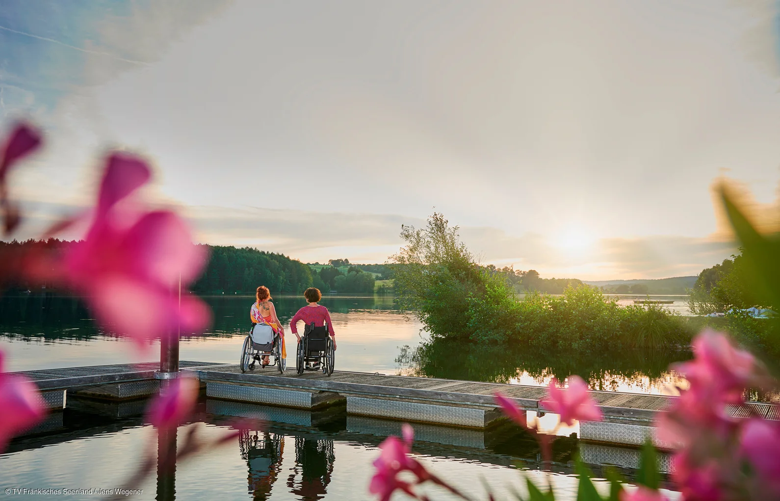 Zwei Personen im Rollstuhl sitzen auf einem Steg am See, Sonnenuntergang und Blumen im Vordergrund.