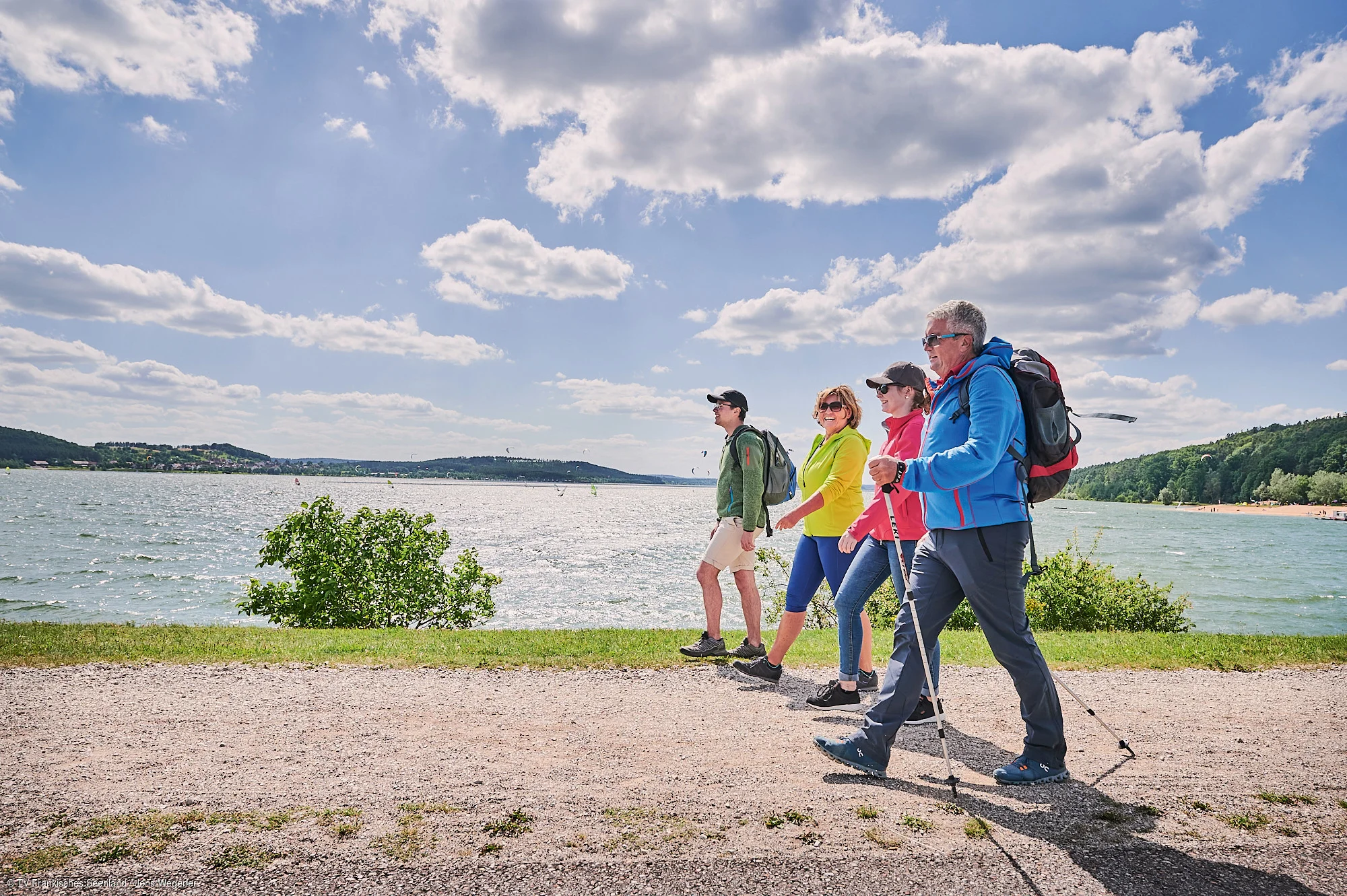 Vier Personen mit Rucksäcken wandern auf einem Weg entlang eines Sees bei bewölktem Himmel.