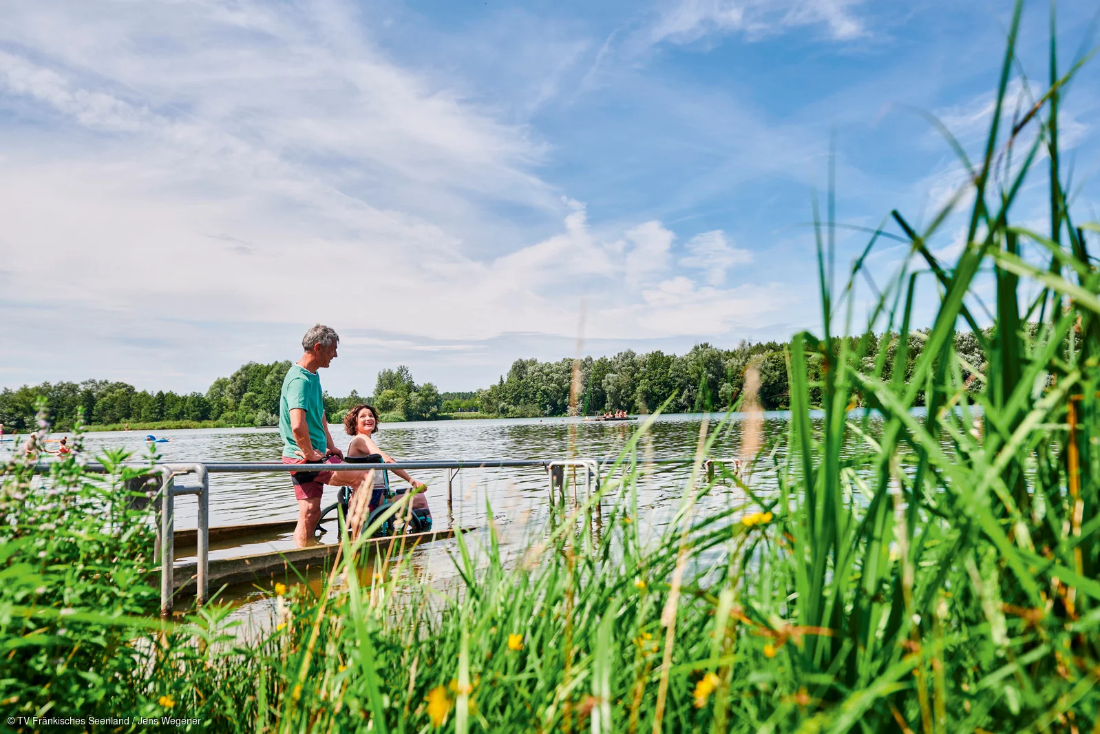 Mann und Frau mit Rollstuhl am Seeufer auf Steg mit Wasser und Bäumen im Hintergrund bei sonnigem Himmel