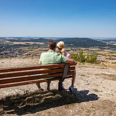 Paar sitzt auf einer Bank auf Hügel mit Blick auf Tal, Felder und Dorf unter blauem Himmel.