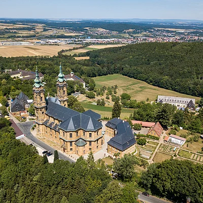 Luftaufnahme eines historischen Klostergebäudes mit Türmen, umgeben von Wald, Feldern und Ortschaften im Hintergrund.