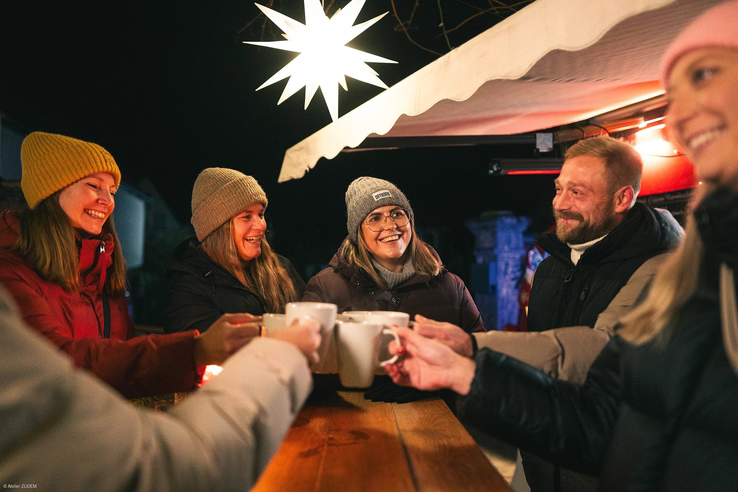 Gruppe von sechs Personen mit Winterkleidung stößt mit Tassen an einem Holztisch unter einem Sternlicht an.