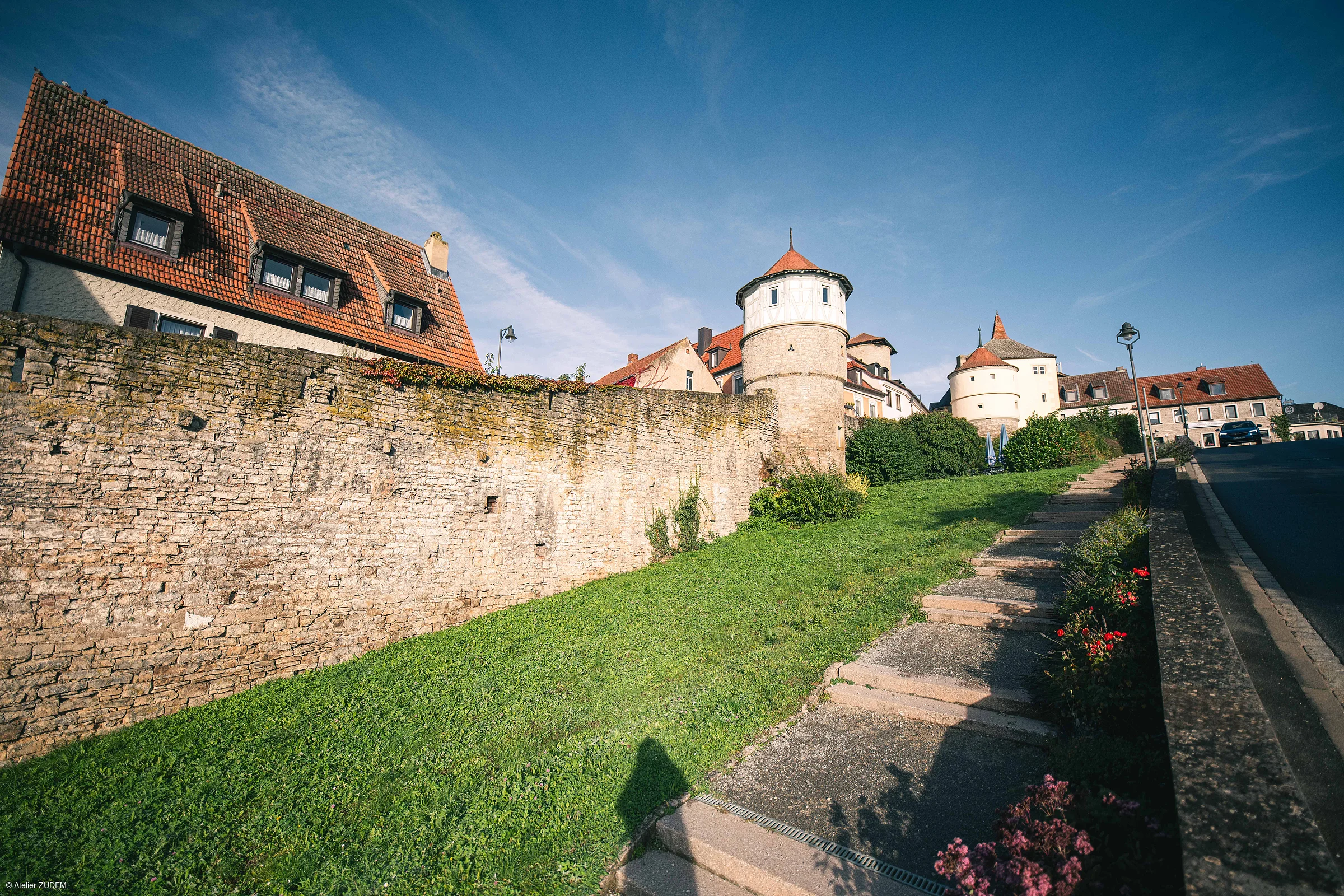 Steinerne Stadtmauer mit zwei runden Türmen, daneben Treppe und Häuser mit roten Dächern unter blauem Himmel.