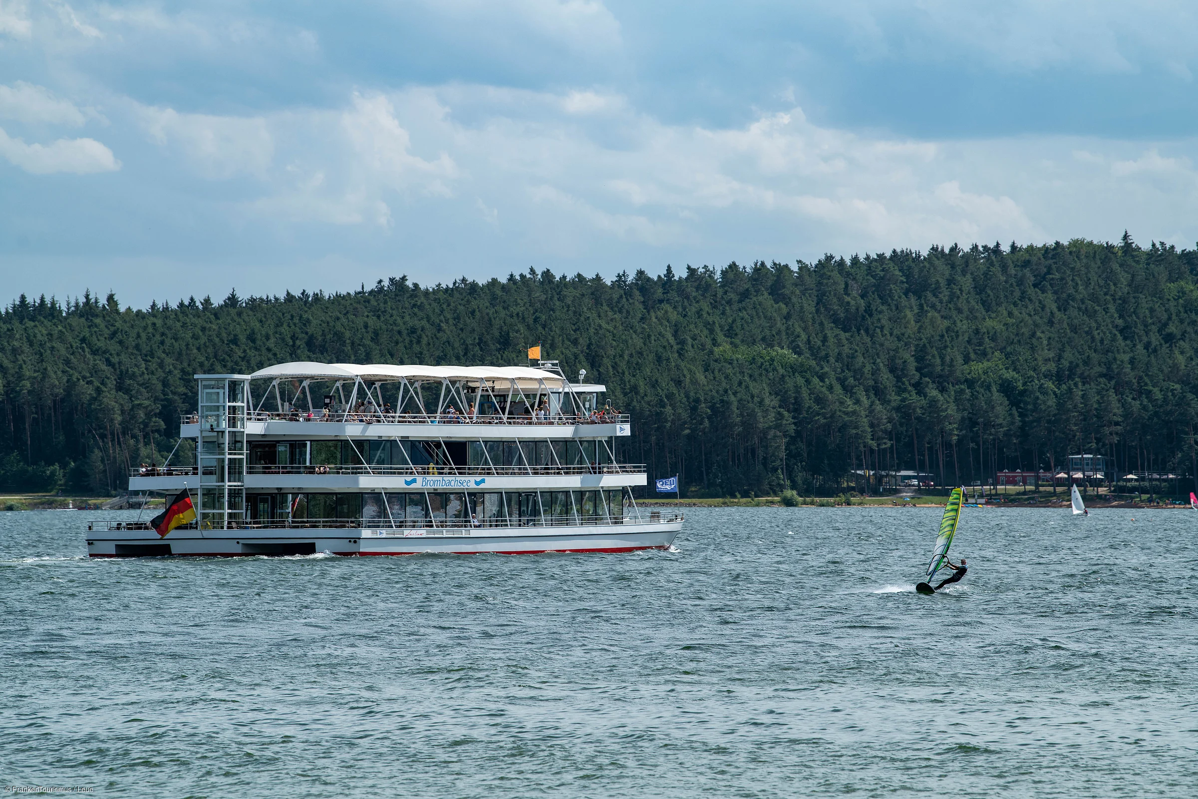 Fahrgastschiff auf See mit Windsegler und Wald im Hintergrund unter bewölktem Himmel.