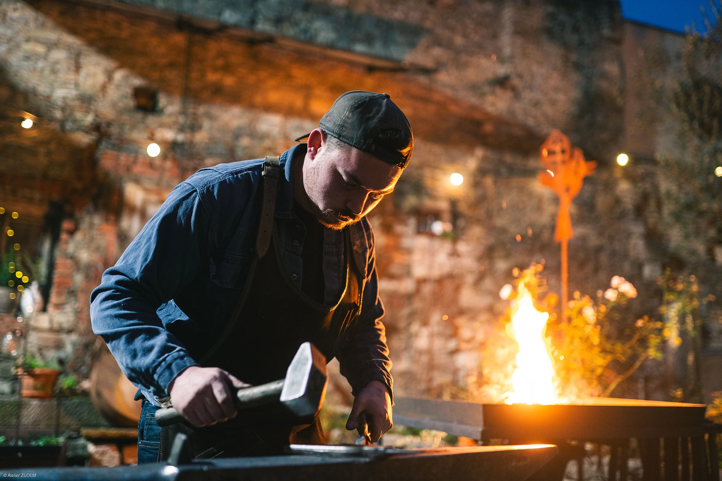 Mann mit Baseballkappe arbeitet abends mit Hammer an Amboss vor Feuerstelle im Freien.