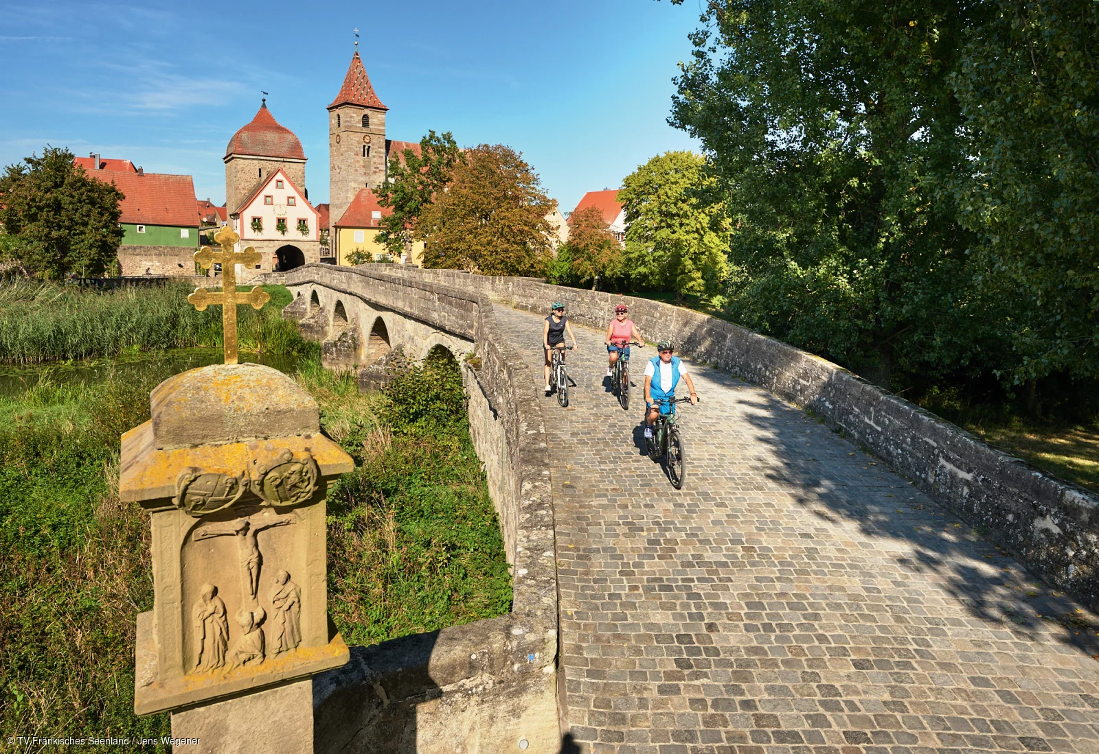 Drei Radfahrer fahren auf einer gepflasterten Steinbrücke mit historischen Gebäuden und Bäumen im Hintergrund.