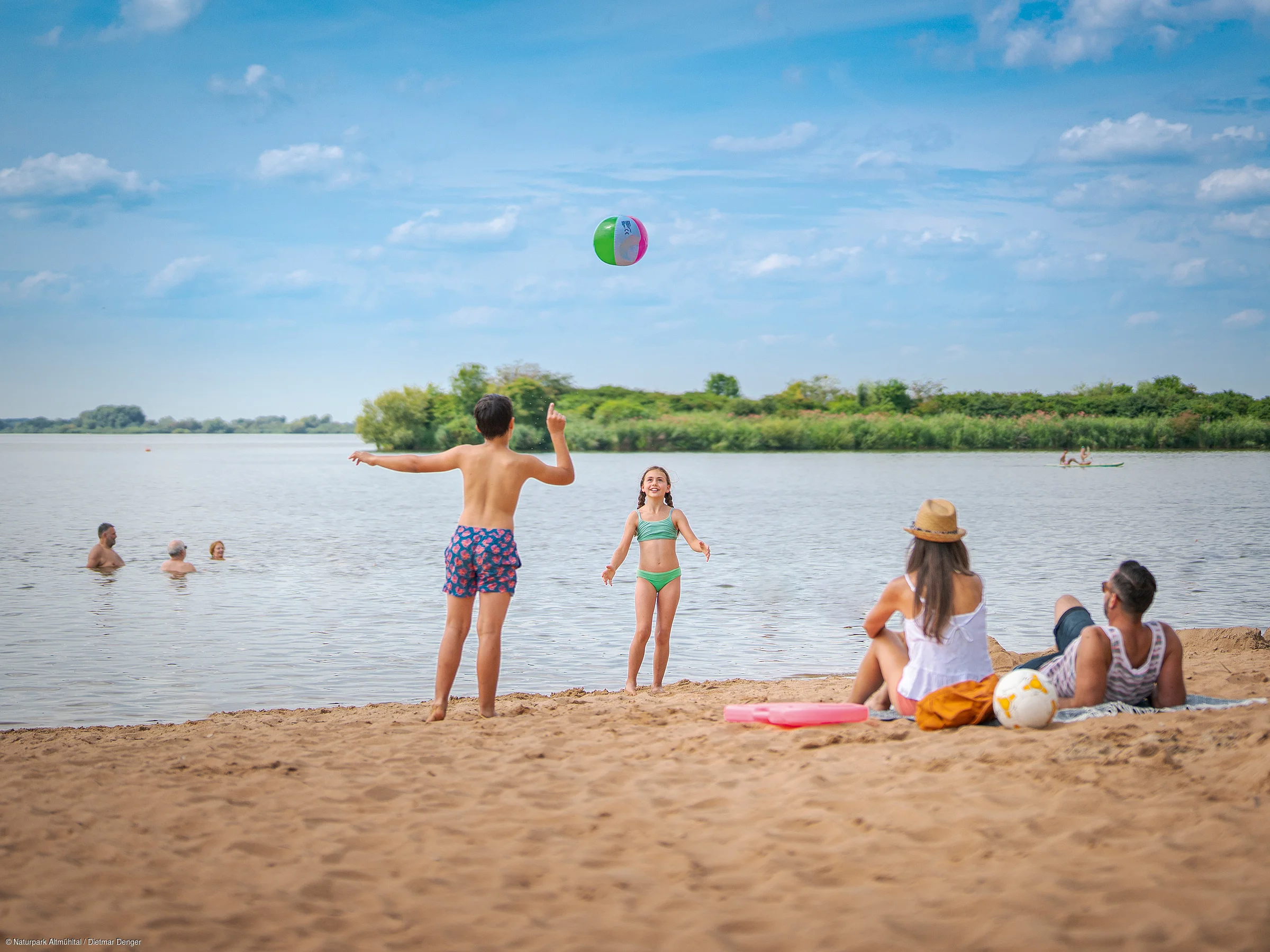 Personen spielen mit einem Ball am Strand, während andere im Wasser schwimmen. Im Hintergrund sind Bäume zu sehen.