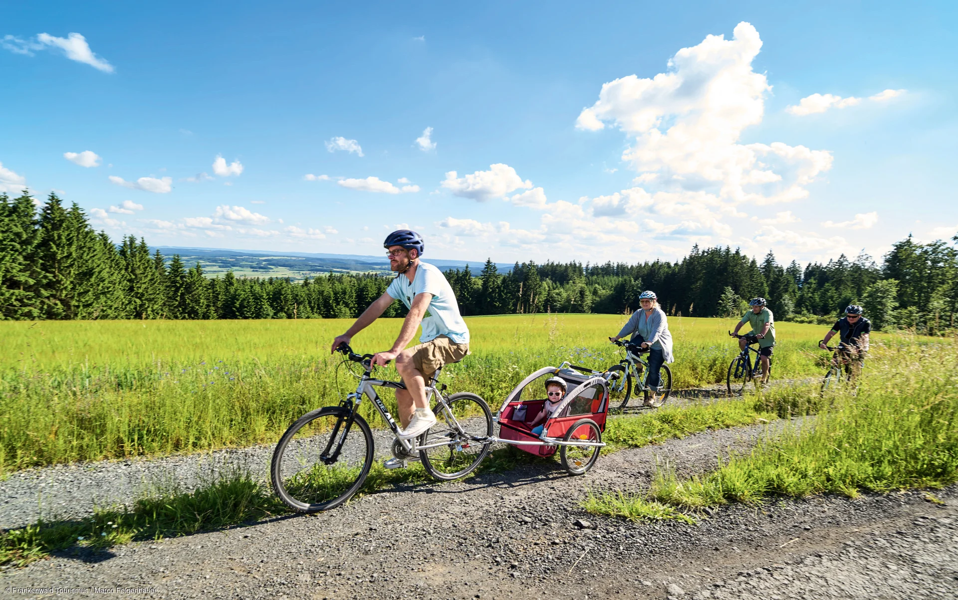 Vier Radfahrer auf einem Feldweg, einer zieht einen Kinderanhänger mit einem Kind, umgeben von Wiesen und Wald.