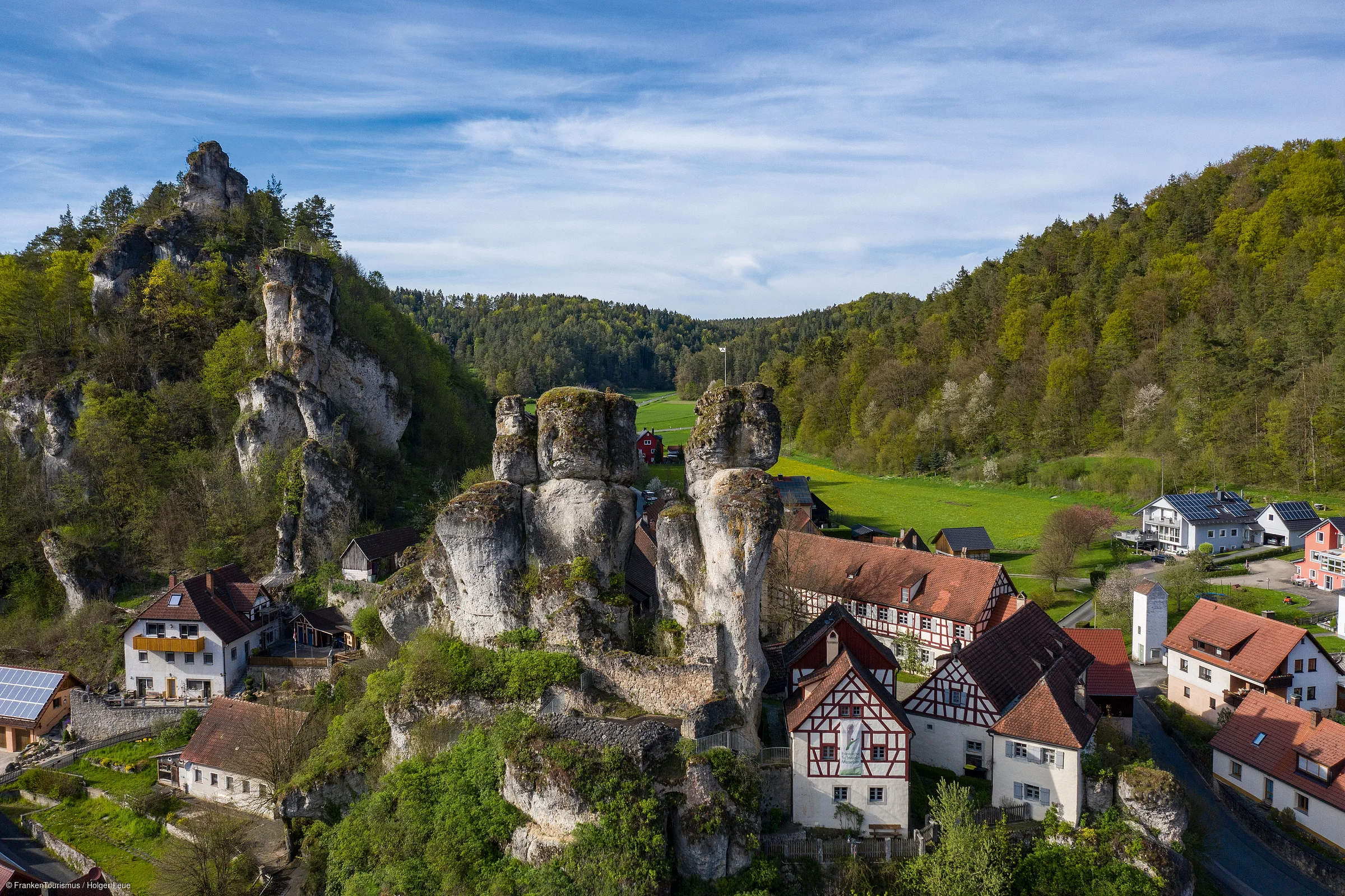 Dorf mit Fachwerkhäusern und Felsen umgeben von bewaldeten Hügeln unter blauem Himmel