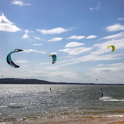 Kitesurfer mit bunten Lenkdrachen auf einem See bei leicht bewölktem Himmel und Sandstrand im Vordergrund.
