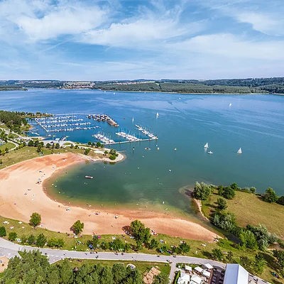 Luftaufnahme eines Sandstrands mit Liegewiese, Bootshafen und Segelbooten auf einem großen See unter blauem Himmel.