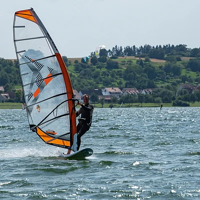 Mann beim Windsurfen auf einem See mit Ufer und Häusern im Hintergrund bei Tageslicht.