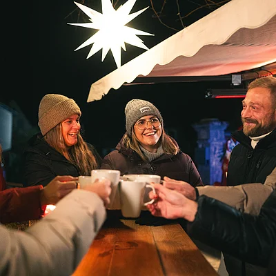 Gruppe von sechs Personen mit Winterkleidung stößt mit Tassen an einem Holztisch unter einem Sternlicht an.