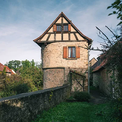 Steinernes Fachwerkhaus mit roten Fensterläden und umliegender grüner Vegetation bei klarem Himmel.