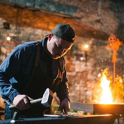 Mann mit Baseballkappe arbeitet abends mit Hammer an Amboss vor Feuerstelle im Freien.