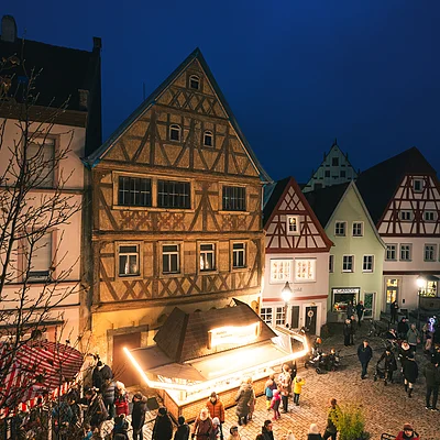 Abendaufnahme eines beleuchteten Marktplatzes mit Fachwerkhäusern und Menschen in Winterkleidung.