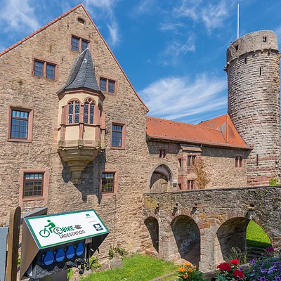 Steinernes Schloss mit Turm, Brücke und E-Bike Ladestation bei sonnigem Himmel und Blumen im Vordergrund.