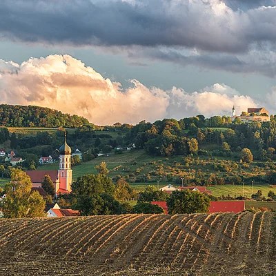 Landschaft mit abgeerntetem Feld, Dorf mit Kirche und bewaldeten Hügeln unter bewölktem Himmel.