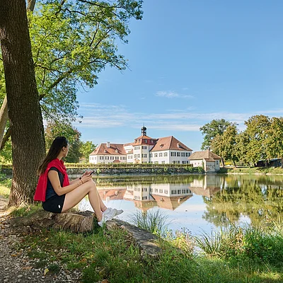 Frau sitzt an einem Seeufer auf einem Baumstamm, vor ihr spiegelt sich ein Schloss im Wasser.