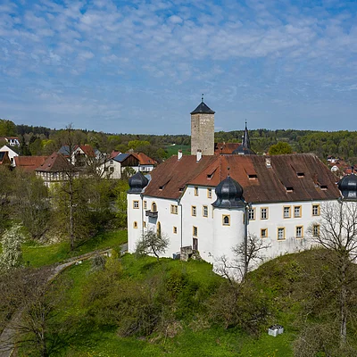 Weißes Schloss mit Türmen auf einem Hügel vor Dorf und Wald unter blauem Himmel mit Wolken