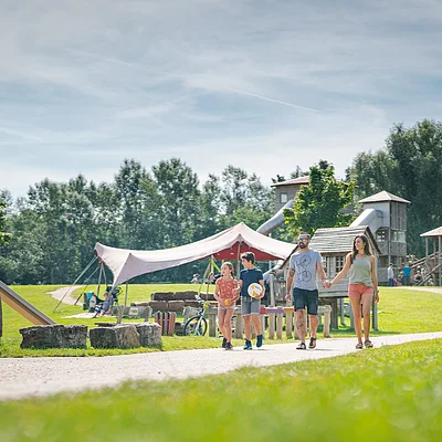 Familie mit zwei Kindern geht auf Weg in einem Park mit Spielplatz und Zelt bei sonnigem Wetter spazieren.