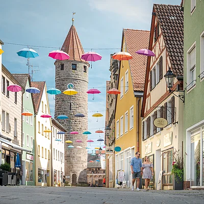 Straße mit bunten Regenschirmen über Kopf und historischem Steinturm im Hintergrund, zwei Personen gehen.