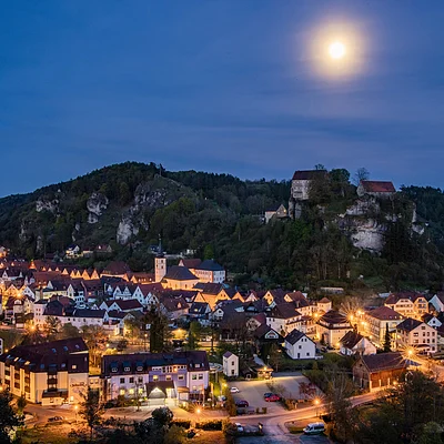 Nächtliche Stadtansicht mit beleuchteten Häusern, Hügeln und Vollmond am klaren Himmel