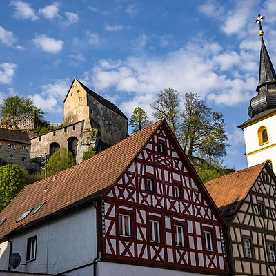 Fachwerkhäuser und Kirchturm vor bewaldetem Hügel mit Burg unter blauem Himmel und Wolken.